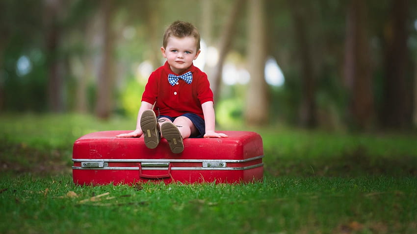 Netter kleiner Junge sitzt auf dem Koffer und trägt ein rotes und blaues Kleid auf unscharfem Bokeh-Hintergrund HD-Hintergrundbild