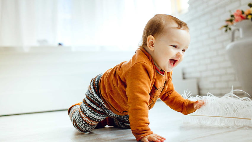 Усмихнато сладко бебе пълзи по пода, облечено в оранжев пуловер, играейки си с бяло перо Сладко HD тапет