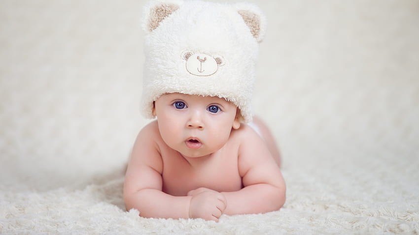 Słodkie dziecko malucha z niebieskimi oczami leży na białej tkaninie na sobie wełnianą czapkę z dzianiny futrzanej śliczną Tapeta HD