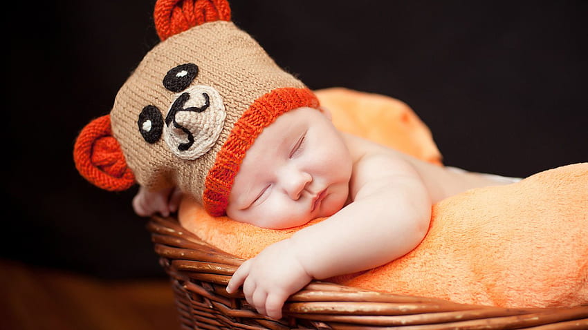 Mignon bébé tout-petit dort à l'intérieur d'un panier en bois sur une serviette orange douce et légère portant un bonnet tricoté en laine sur fond sombre mignon Fond d'écran HD