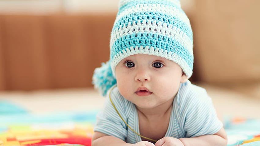 Nettes schönes Kind Baby trägt blaues weißes Kleid und wollene gestrickte Mütze, die auf buntem Tuch im unscharfen Hintergrund liegt nett HD-Hintergrundbild