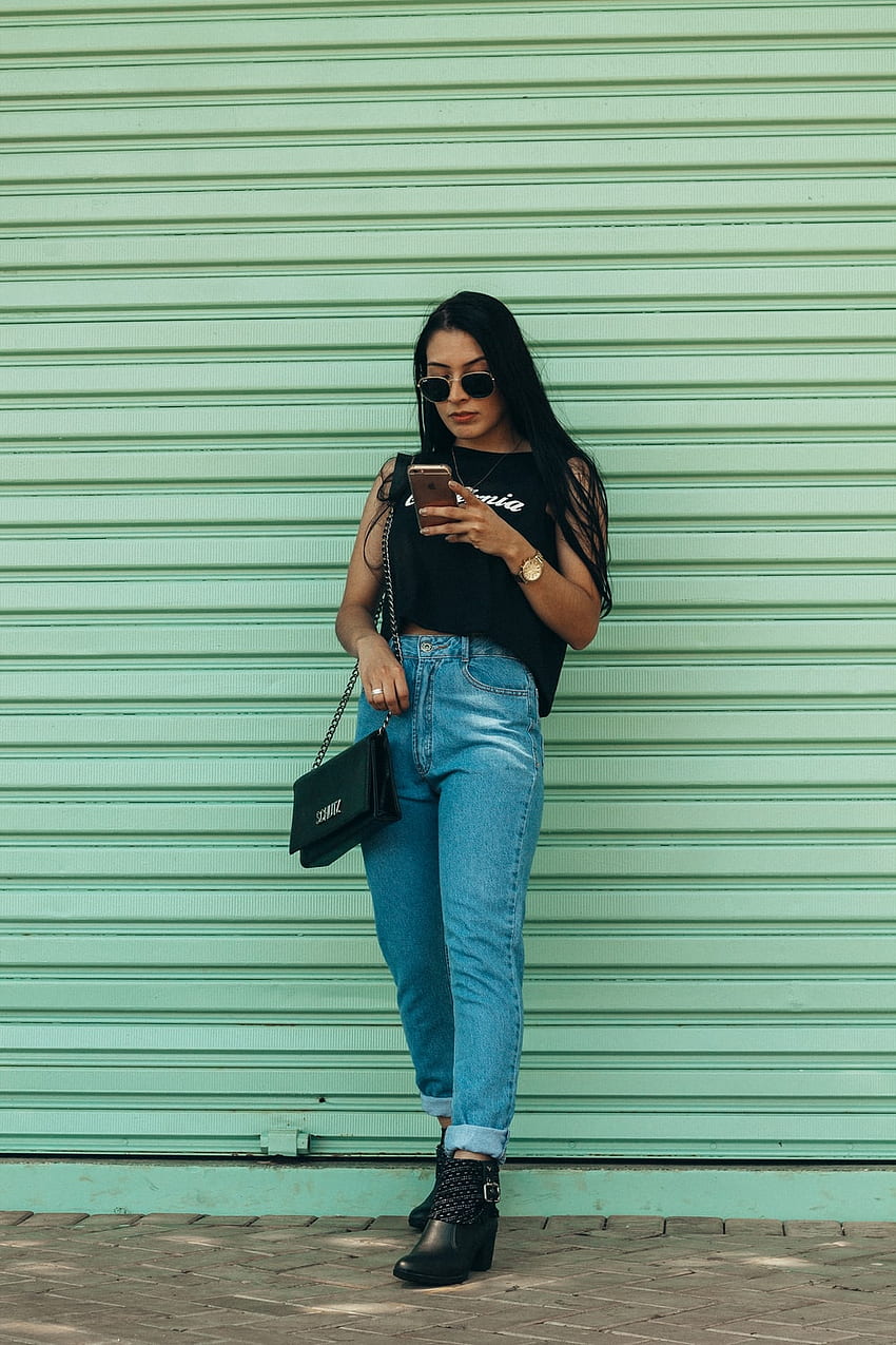 屋外でスマートフォンを保持している黒のノースリーブ トップの女性 – ジーンズ、緑、黒のスマートフォン HD電話の壁紙