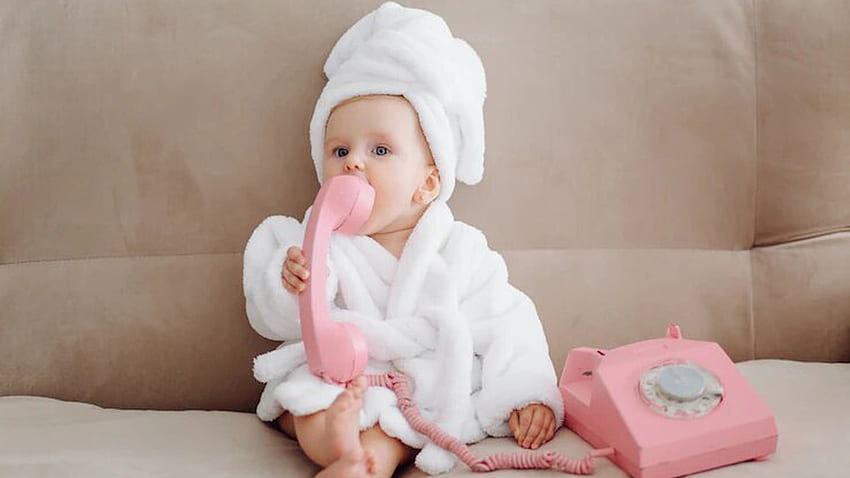 Сладко момиче Бебе носи бял халат, седи на дивана и има розов телефон Телефонна слушалка на уста Сладко HD тапет