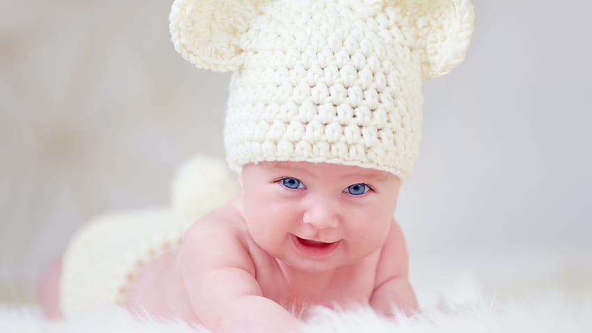 Sonriente ojos azules lindo bebé niño pequeño está acostado sobre un paño de piel blanca con gorro de punto de lana lindo fondo de pantalla