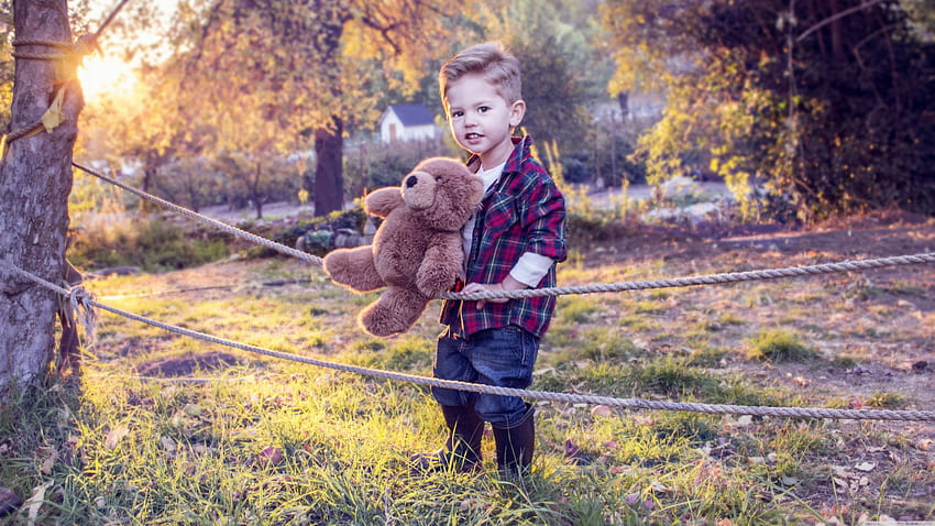 Lindo niño pequeño con juguete de peluche está de pie sobre la hierba en el del jardín vistiendo camisa a cuadros azul rojo y jeans azul lindo fondo de pantalla