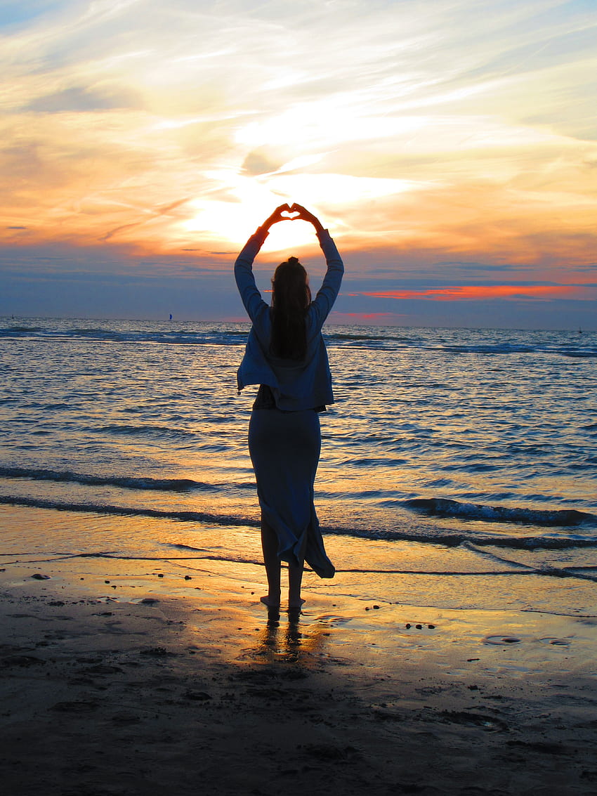 Жена с вдигнати ръце, която прави знак със сърце, докато стои на плажа, на залез сърце HD тапет за телефон